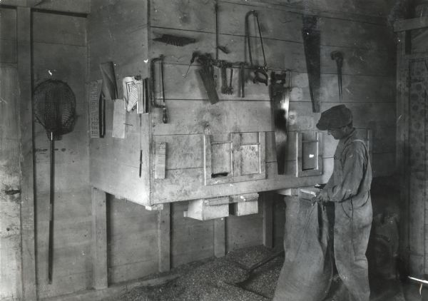 A man wearing a cap and overalls holding a burlap feed bag while standing near feed bins in the poultry house of Professor Holden. Tools are hanging from nails hammered into the wooden walls.