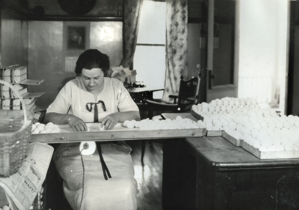 A woman wearing a dress sitting indoors to candle eggs at the Holden Farm. Cartons of eggs are piled on a table to her left, and household furniture is in the background.