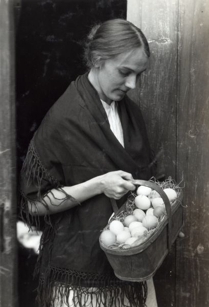 A woman wearing a fringed shawl is standing in the doorway of a barn holding a basket of eggs.