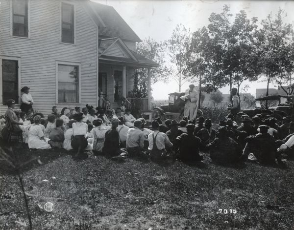 A group of men and women gather around Professor Garrick in the yard and on the porch of a farmhouse to watch a poultry culling demonstration. Additional farm buildings and two automobiles are in the background.