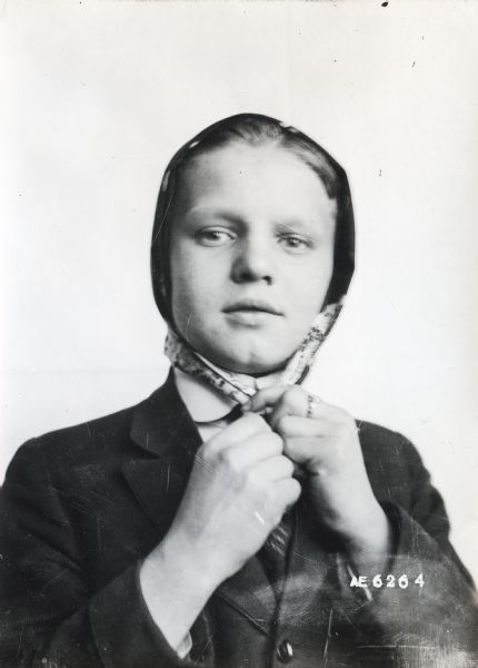 Quarter-length portrait of a woman tying a scarf around her head and knotting it under her chin. The photograph is one of a series demonstrating how to use a scarf as a head covering.