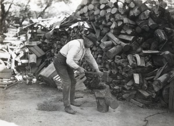 A man wearing a hat and suspenders is using an axe to slaughter a chicken, using a log as a base. A large pile of firewood is behind him.
