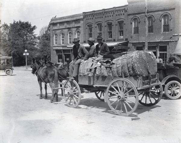 Three men wearing hats are sitting on top of baled cotton loaded in a wagon. The wagon is led by two horses and is traveling down a dirt road in what appears to be a commercial district, with signs on the storefronts in the background reading: "Goodyear Shoe Shop," "Groceries," and "Undertaking."