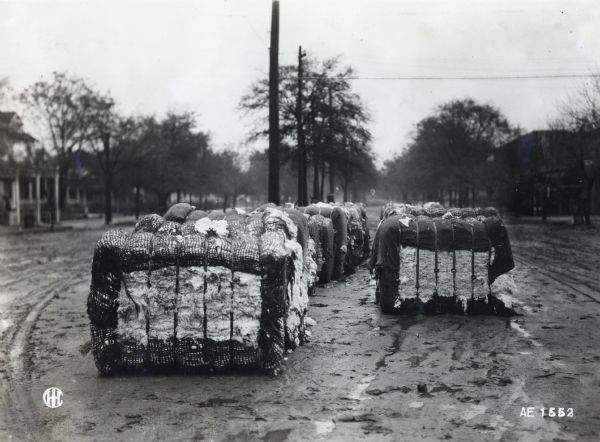 Bales of cotton stored in the middle of a damp dirt road. Houses or storefronts are on both sides of the road in the background.