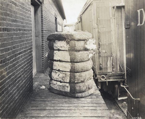 A bundled cotton bale rests on a wooden platform at a loading dock or train depot. A train car on a railroad track is alongside the right of the platform.