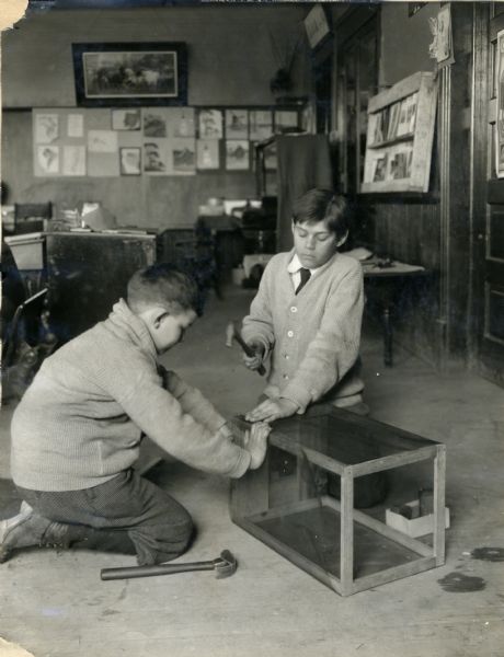 Two boys construct a fly trap. A boy holds mesh in place against a wooden box frame while another boy uses a hammer to nail it into place. They kneel in what appears to be an office or schoolroom, with furniture, and various objects are hanging on the walls in the background.