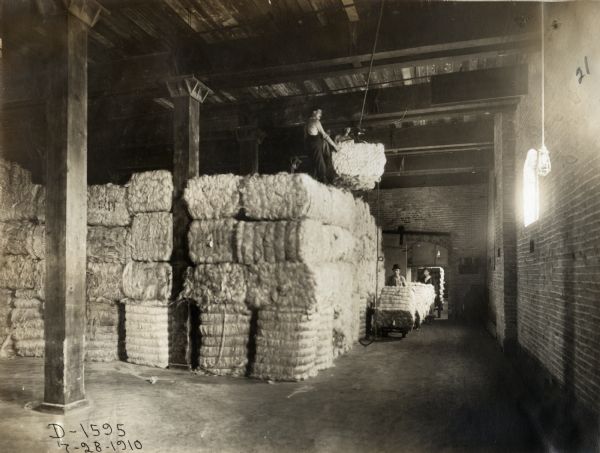 Men use a pulley to raise bales of sisal from the ground of a warehouse to the top of a pile of stacked bales. Others use dollies to wheel in additional bales through an open door. The warehouse is likely on the grounds of the McCormick Twine Mill in Chicago.