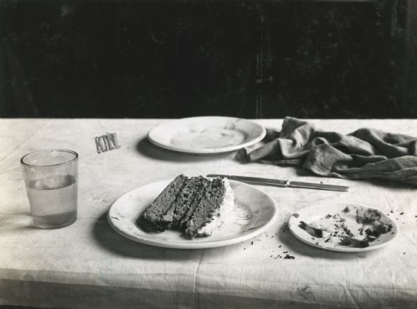 A slice of frosted cake rests on a plate atop a cloth-covered table, along with a glass, knife, additional plates, and a wrinkled cloth napkin.