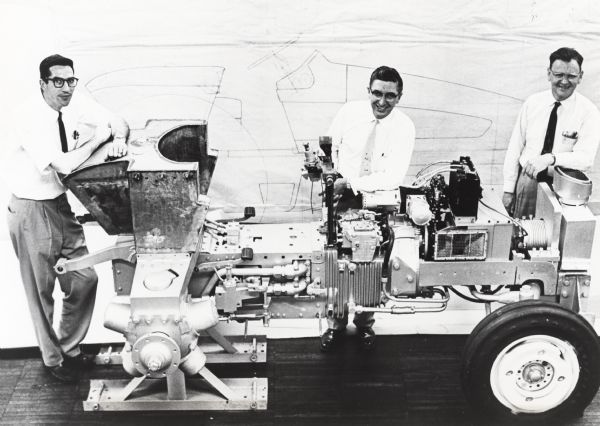 A profile view of Harvester's HT 340 tractor on assembly stand showing a rear mounted fuel tank, front exhaust, and side air intake. Personnel in the group portrait are, left to right: John R. Cromack, test engineer; Carl H. Meile, chief engineer; and Ralph E. Wallace, research engineer. A full scale line drawing of the experimental tractor is on the wall behind the men.