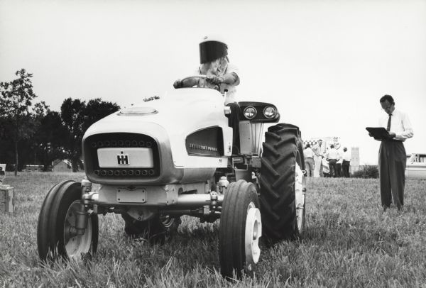 An experimental International HT 340 tractor is being tested by a man wearing a helmet in an outdoor environment. Other men are standing in the background.