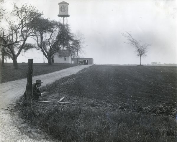 A man crouching in a farm field near a wooden tool, possibly doing irrigation work. A dirt road behind the man leads to several buildings and a water tower marked: "IHC." The site may be one of International Harvester's experimental farms.