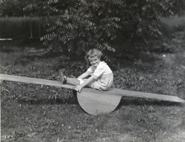 A girl with bobbed hair is wearing a one-piece playsuit, knee socks, and buckled sandals while playing on a seesaw (teeter totter).