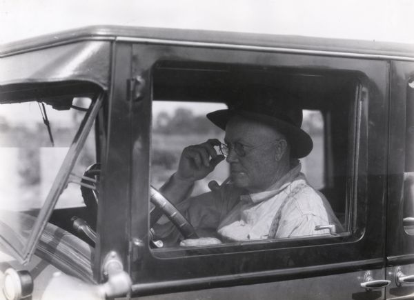 John Esch, sitting behind the wheel of an automobile, adjusting his eyeglasses while smoking a tobacco pipe. Photograph taken for International Harvester's Agricultural Extension Department to illustrate the dangers of driving with impaired eyesight.