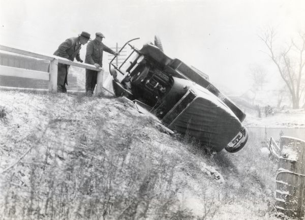 Two men standing by a guard rail along the side of a road to look at a snow-covered automobile resting on its side.
