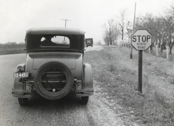 Rear view of a man wearing a wide-brimmed hat stopping an automobile at a stop sign on a dirt road as a truck approaches from the opposite direction.