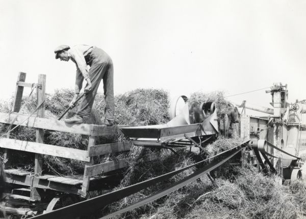 A farmer is standing on the edge of a wooden wagon pitching hay into a running thresher, demonstrating an unsafe operating position.