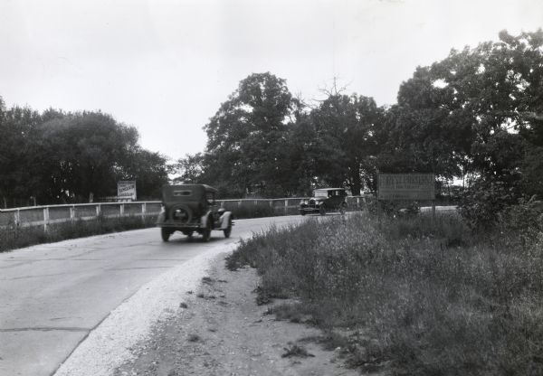 Automobiles approaching a turn in a paved road with billboards on either side. Photograph taken for International Harvester's Agricultural Extension Department to illustrate safe driving techniques.