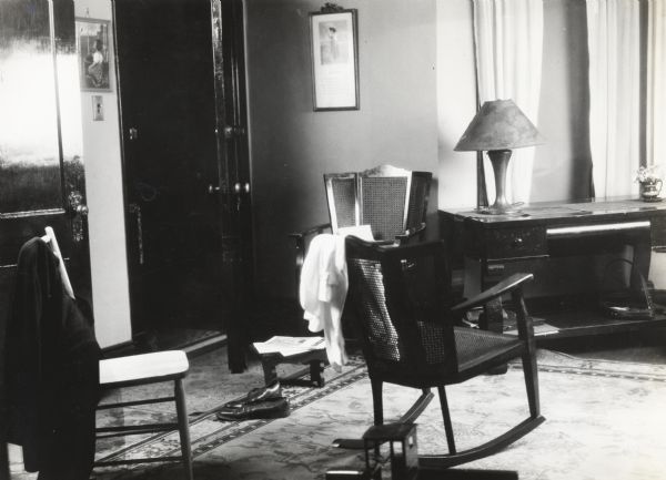 Interior view of a farmhouse, taken for International Harvester's Agricultural Extension Department and used to illustrate the dangers of a cluttered room. Several chairs, a pair of shoes, and a step-stool with magazines piled on it block the way to an opened door.