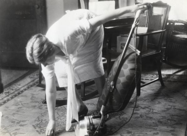 A woman stooping to remove an electrical cord from in front of a vacuum cleaner.