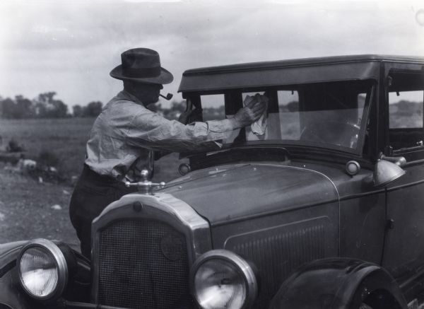 A man wearing a hat and smoking a tobacco pipe using a rag to wipe the windshield of an automobile.