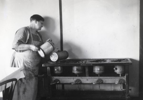 A woman wearing an apron pouring oil into a hot stove at the International Harvester Hinsdale experimental farm, illustrating a dangerous scenario for International Harvester's Agricultural Extension Department.