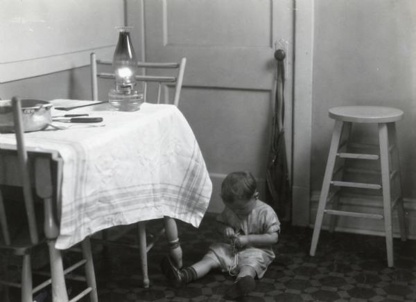 A child sitting on the floor of a farmhouse kitchen near a cloth-covered table. A lit oil lamp, pot, and utensils are sitting on the table, illustrating the danger of leaving children unattended around dangerous objects on a farm.