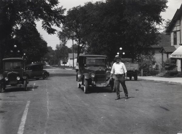 A man jaywalking across a street in front of a moving automobile. The road is lined with houses and commercial buildings, and additional automobiles are parked alongside the curb.