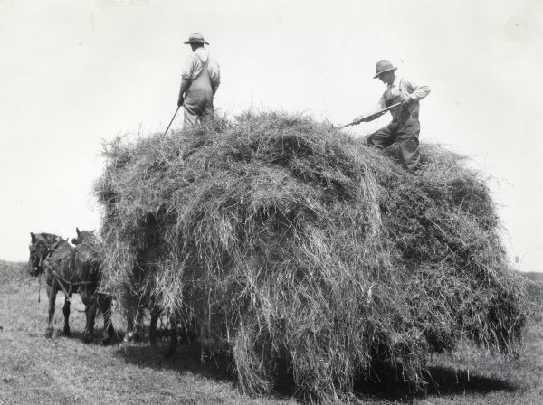 Two men holding pitchforks stand near the edge of a horse-drawn wagon piled with hay.