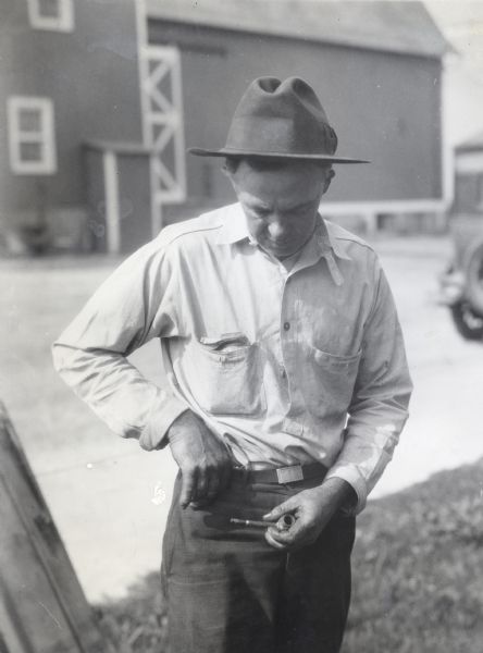 A man reaching for loose matches carried in his pocket while holding a tobacco pipe in his other hand. The photograph was taken on International Harvester's Hinsdale experimental farm, and an automobile and barn are in the background.