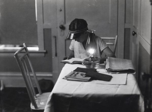 A woman sitting at a kitchen table wearing a visor, leaning over a book while a lit oil lamp is nearby. Also on the table are a newspaper, additional books, and papers.