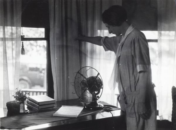 A woman moves a curtain out of the way of an electrical fan sitting on a desk. Near the fan are some books, a floral arrangement, and a notepad. Outdoors, visible through the window, is a parked automobile.