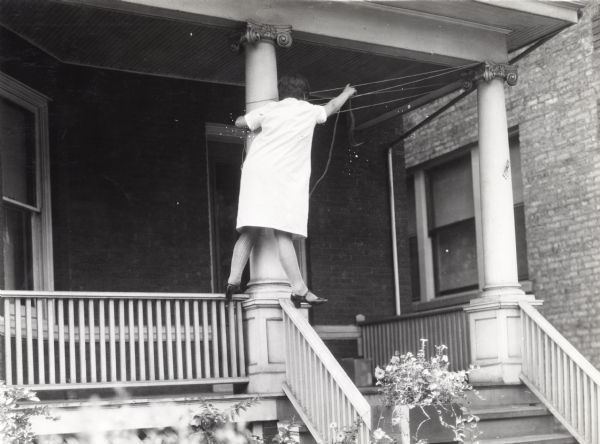 A woman wearing a house dress, stockings, and heeled shoes standing on the porch railing of a brick house to hang what appears to be clothesline from porch pillars.
