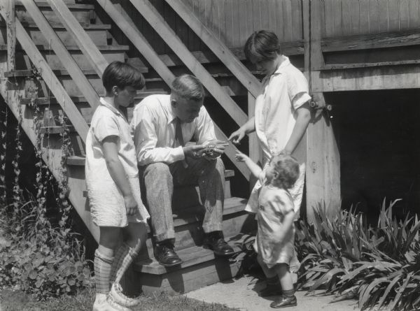 A man sits on wooden outdoor stairs holding a firearm, while three children standing around him.