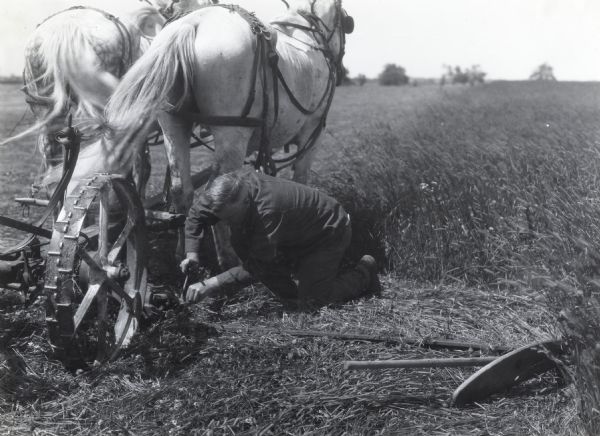 A man is kneeling on the ground beside a mower hitched to two horses while performing repairs. An agricultural tool is lying on the ground beside him.