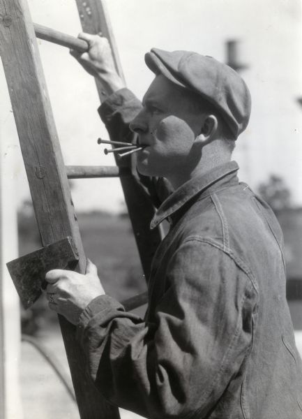 A man wearing a hat and one-piece work suit climbing a wooden ladder while holding nails in his mouth. He appears to be holding an axe in his left hand.
