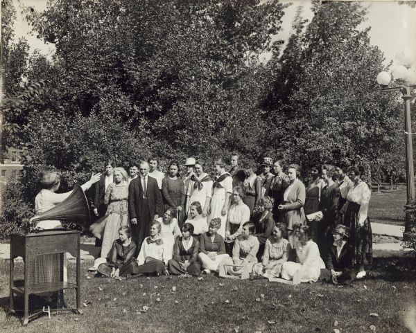 Group of women and man receiving musical instruction outdoors from a teacher using a phonograph. Some of the group is standing, and the rest are sitting on the lawn.