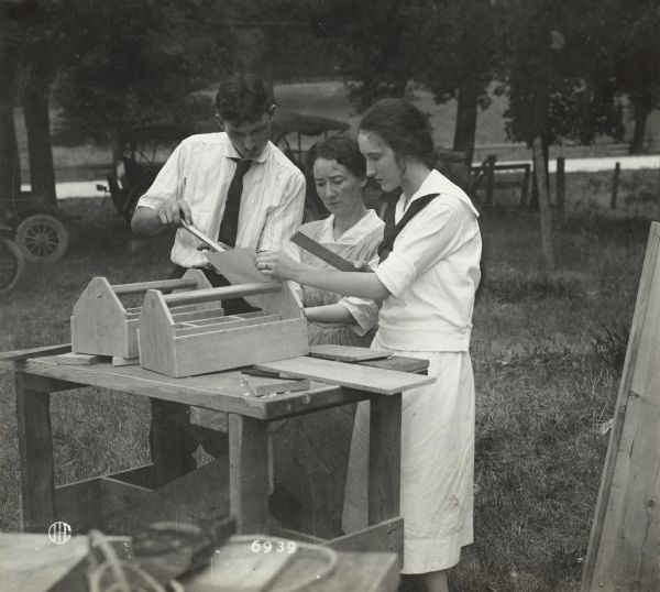 Two women and one man, probably rural school teachers, are standing outdoors near a work bench, reviewing the blueprints to construct a nail box.
