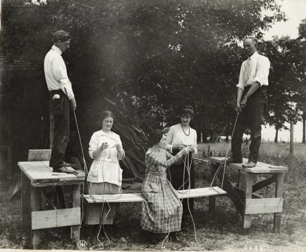 Three women tying rope. One woman is sitting on a wooden plank suspended on both ends by rope held by two men standing on top of workbenches. The men and women are likely rural school teachers.