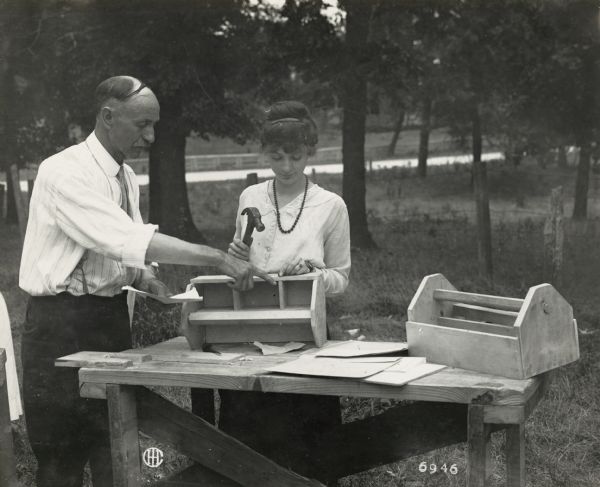 A woman, probably a teacher, is standing at a work bench outdoors nailing together a nail box. Her instructor, identified by original caption as Mr. Shepherd, Country Life Director of Cook county, points out an error.