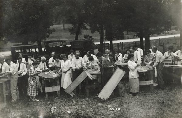 A large group of men and women, probably teachers, stand outside at work benches assembling wood products, which include nail boxes and benches.