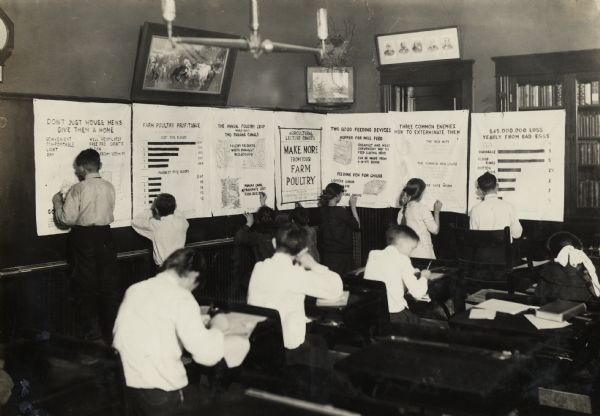 Young students making stenciled charts with teacher J.W. Page at the Morton Grove School. Original caption reads: "Another view of a school making junior charts from the stenciled patterns furnished by the IHC Agricultural Extension Department."