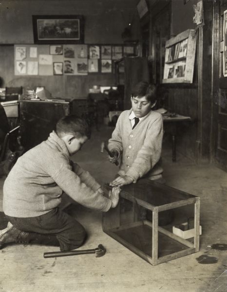 Two young boys on the floor of a room, probably a classroom, nailing mesh screening to the frame of their fly trap.