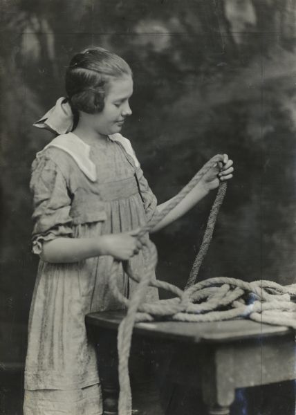 Girl standing at work bench with a length of rope. Original caption reads: "Cleo Holt spliced her father's hay rope."