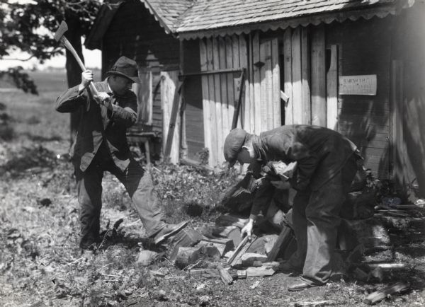 Two men chop wood beside a wooden shed at International Harvester's Hinsdale experimental farm. One man raises an axe while steadying a piece of wood with his foot, and another man stoops to gather chopped pieces. The sign on the shed behind the men reads: "Smoking Absolutely Prohibited."