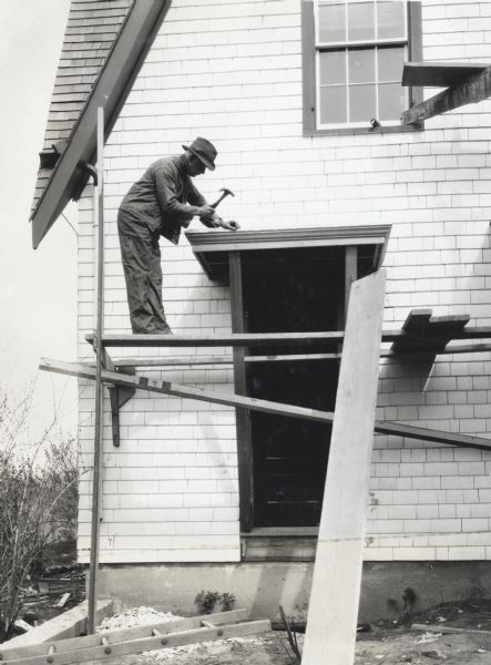 A man stands on makeshift scaffolding while hammering a nail into a door awning. The photograph was taken for International Harvester's Agricultural Extension Department to illustrate hazards found on the farm.