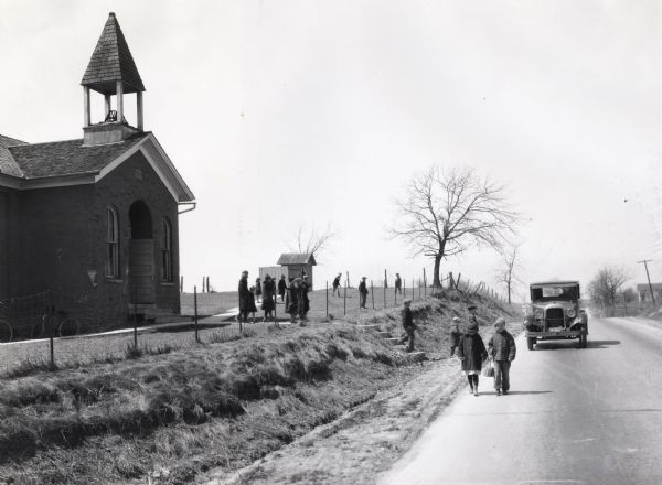 Children walking along the side of a road in front of a one-room schoolhouse as an automobile approaches from behind. Other children are playing in the schoolyard and several bicycles are leaning against the school building.