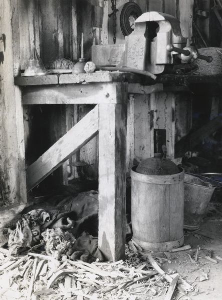 Interior view of a unkempt work space inside a shed or barn. There are wood shavings, dirty rags, and gloves lying beneath the work bench.