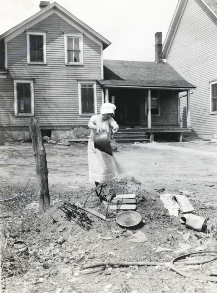 A woman wearing a dress and bonnet empties a pail of ashes into a refuse pile in the front yard of a farmhouse.