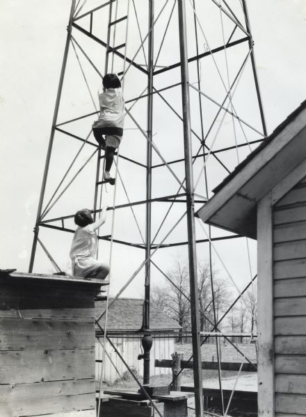 Two girls climb the base of a windmill surrounded by farm buildings.