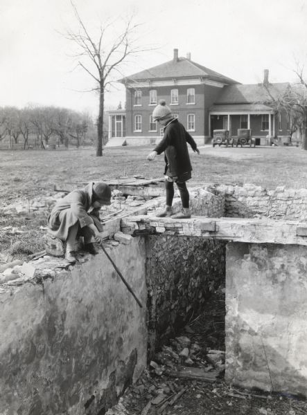 Two children playing on the stone foundation of a basement which appears to have been damaged by fire. A farmhouse with two automobiles parked nearby are in the background.